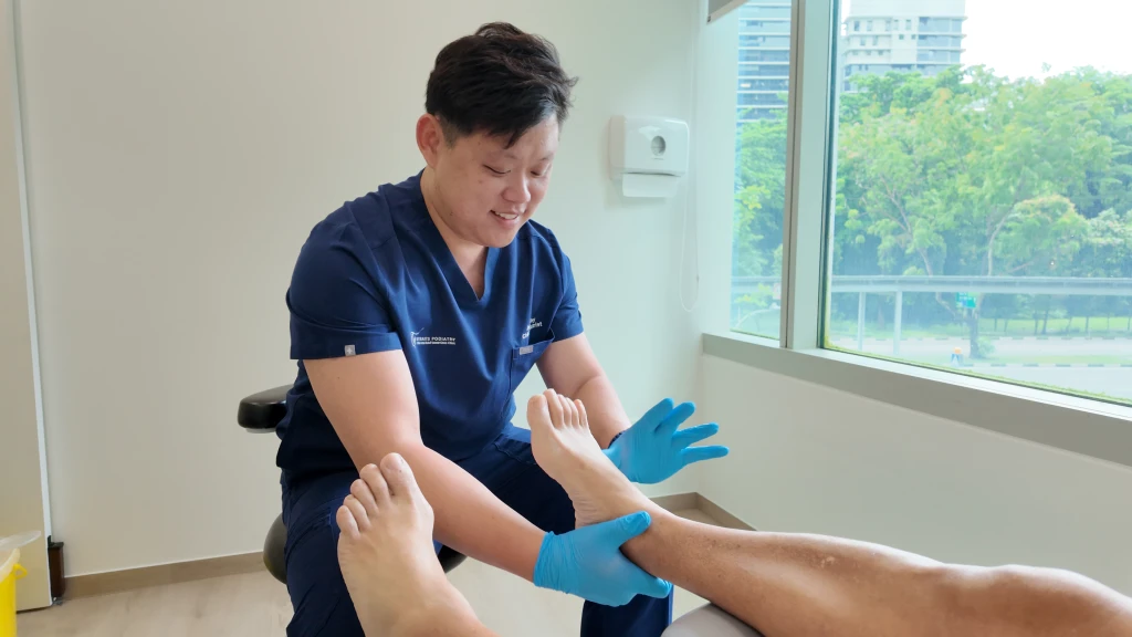 A podiatrist at Straits Podiatry wearing gloves examining a patient's bare foot and lower leg during an Achilles tendonitis assessment.