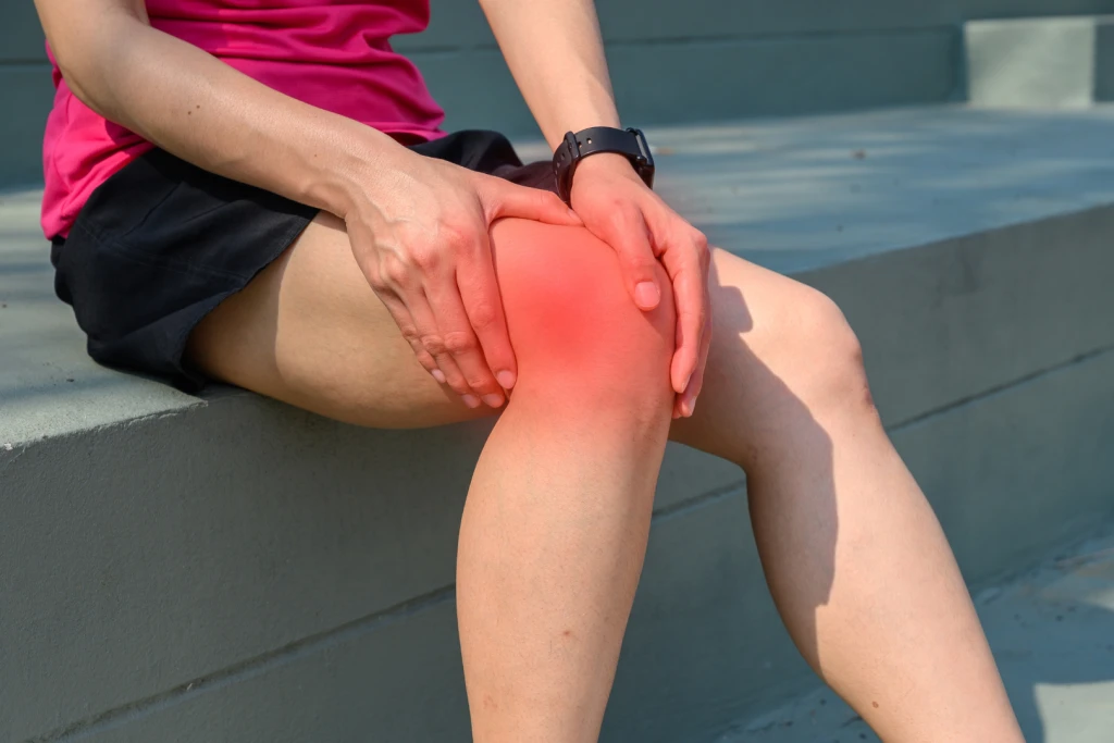 A person in activewear sitting on outdoor steps and gripping the outer side of the knee, highlighted in red, the pain location typical of iliotibial band syndrome.