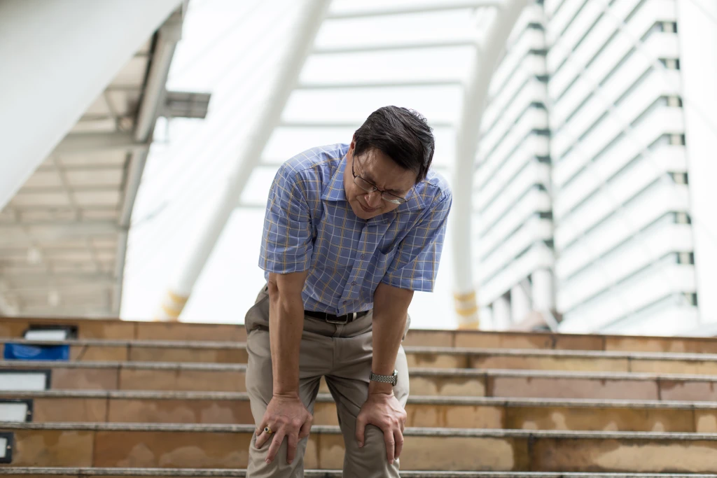 A person in a checked shirt pauses on outdoor stairs, bending forward with both hands gripping the knees, the stair descent that commonly aggravates patellar tendonitis.