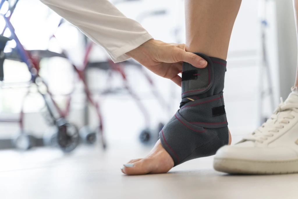 A hand in a white sleeve fastening a grey fabric brace around a standing person's ankle in a clinic, the external support used when managing chronic ankle instability