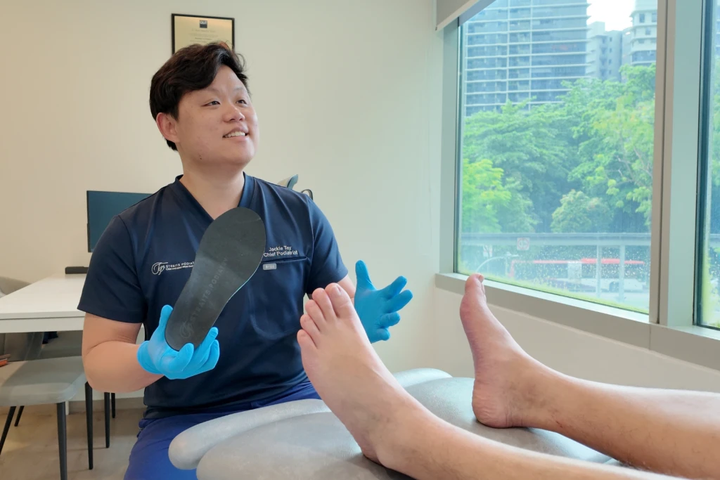 A podiatrist in navy scrubs holds a black foot orthotic while talking with a patient whose bare feet rest on the couch, the offloading approach used for rheumatoid arthritis in feet.