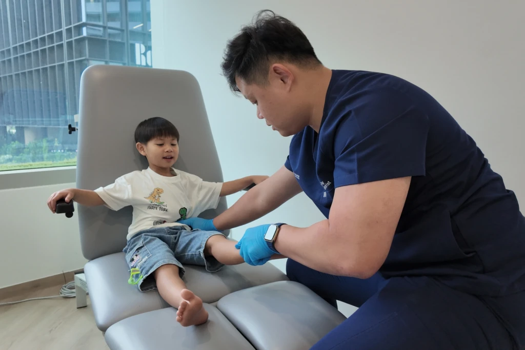 A podiatrist in navy scrubs and blue gloves examines a young child's lower leg and ankle in a clinic chair, part of assessing osteochondrosis