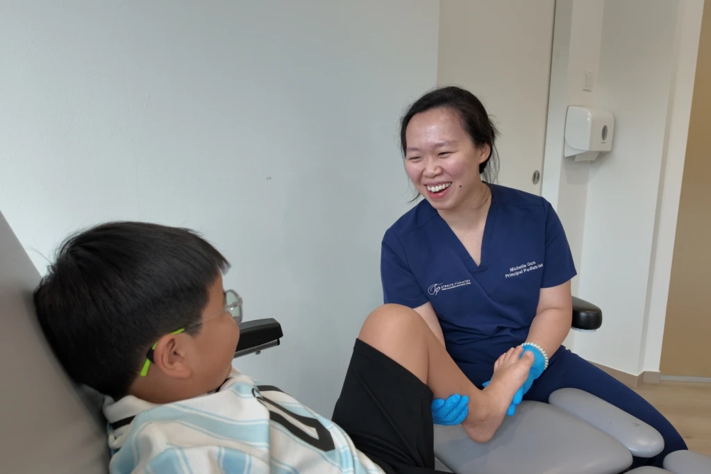 A podiatrist in navy scrubs holds a spiky therapy ball against a smiling child's foot in a treatment chair during a paediatric assessment for Osgood-Schlatter disease.