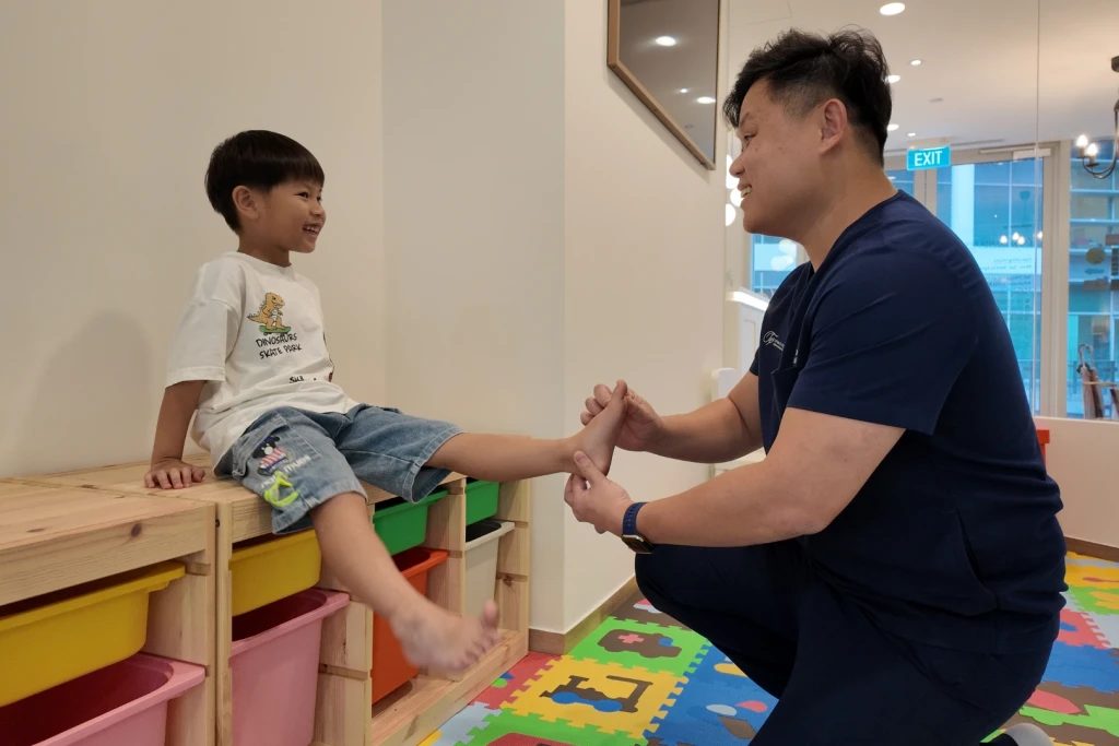 A podiatrist crouches to hold and flex a smiling child's bare foot in a clinic play area, a range of movement check used when assessing osteochondrosis