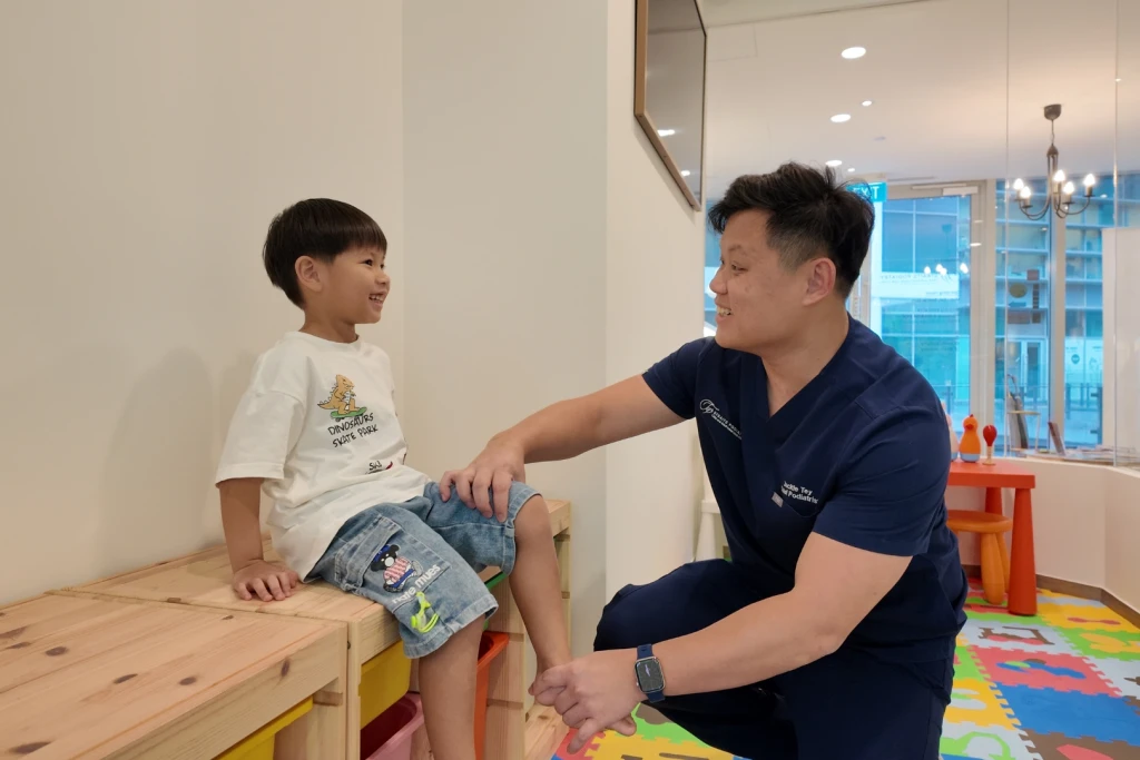 A podiatrist crouching on a colourful play mat holds a smiling child's knee and shin in the clinic play corner, the region involved in Osgood-Schlatter disease.