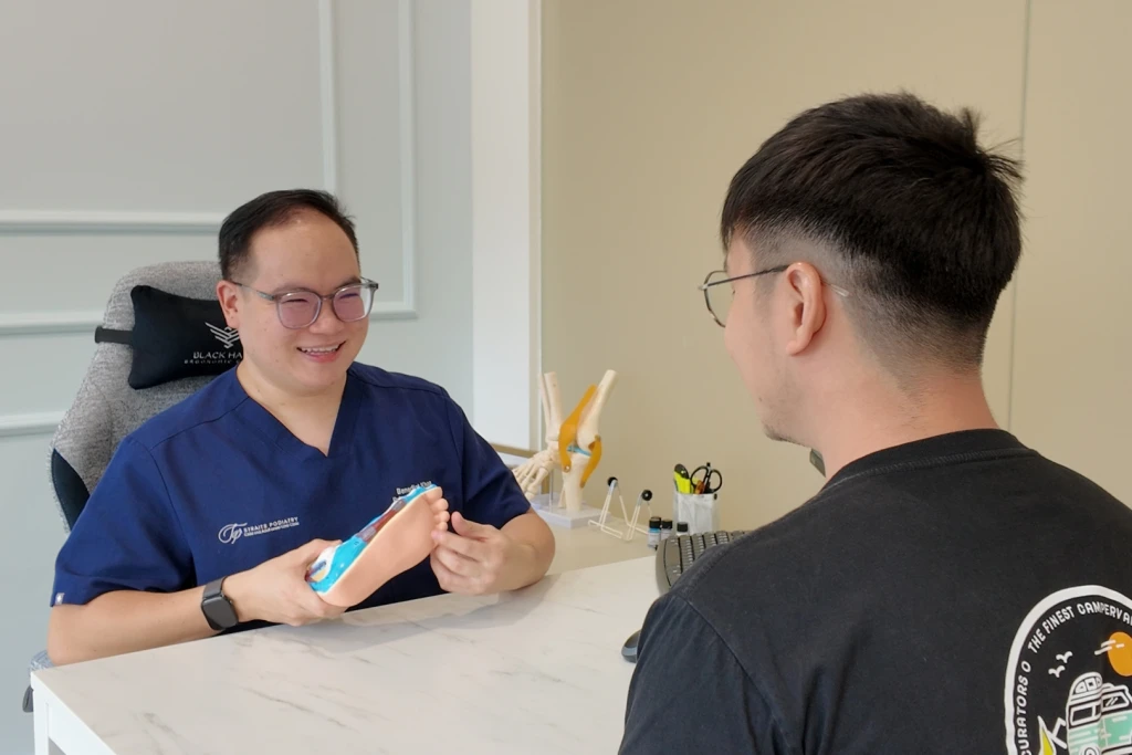 面带微笑的足科医师手持足部解剖模型并指向前足部位,向坐在咨询台对面的患者进行讲解。