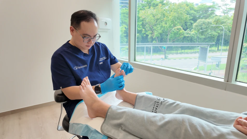 A podiatrist in navy scrubs and blue gloves works with a small blade on a patient's toe beside a clinic window, the careful paring used to manage corns