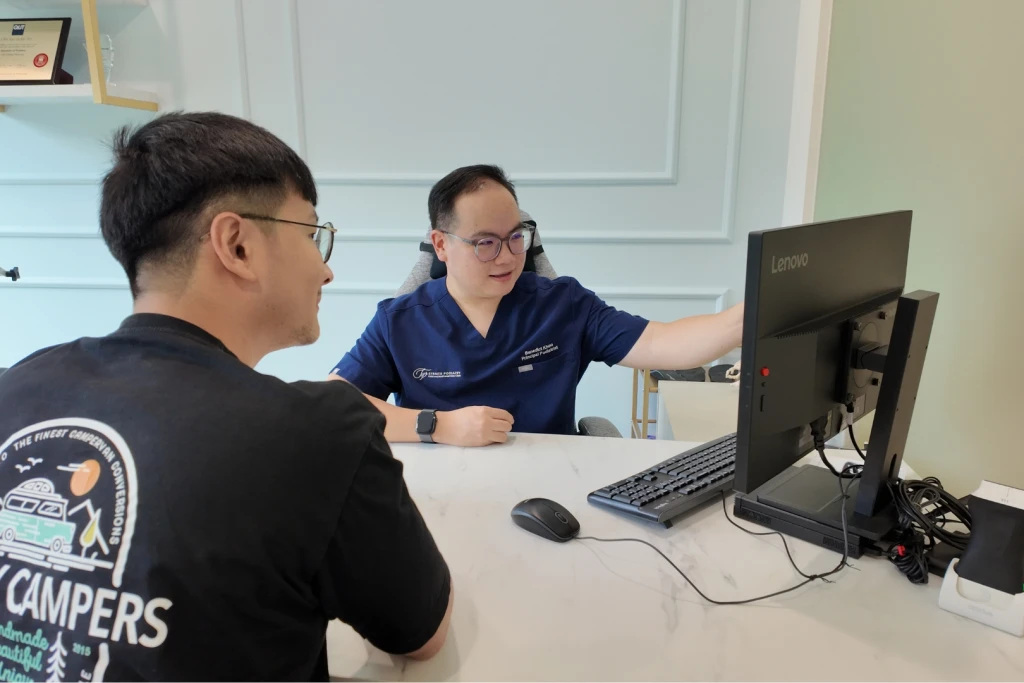 A podiatrist in navy scrubs turning a clinic monitor towards a patient at the desk to go through findings, the review that follows a shin splints assessment.