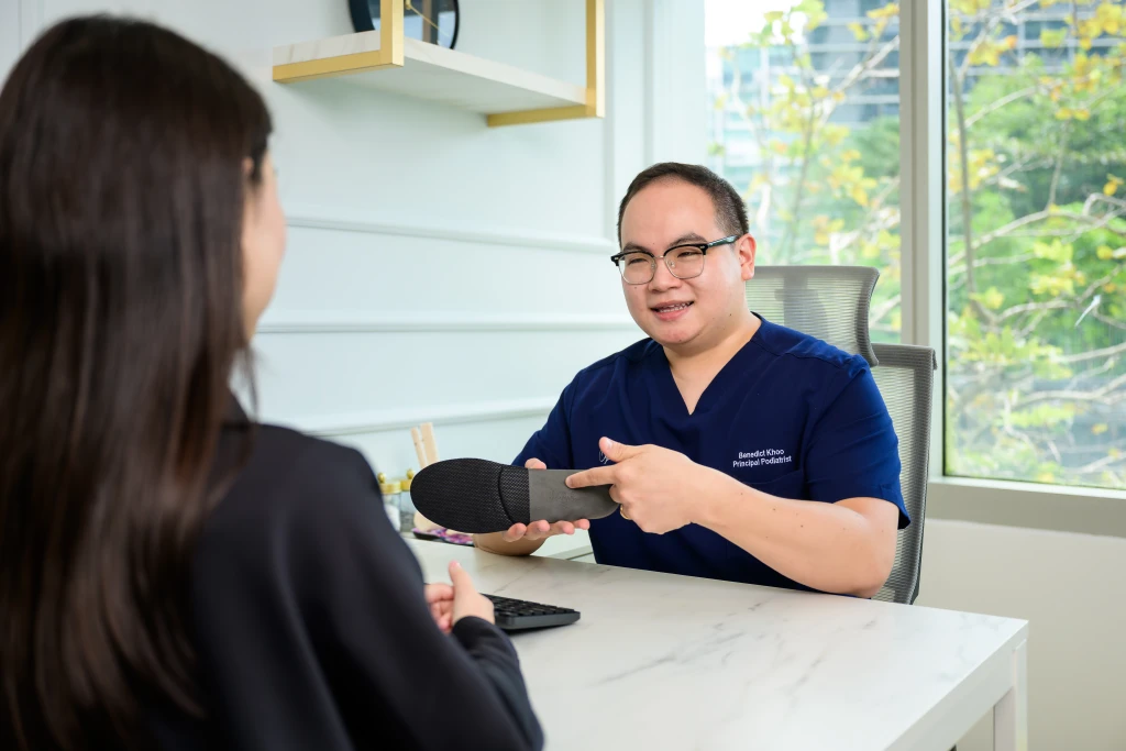 A podiatrist in navy scrubs holds up a black custom orthotic and points at its shell while explaining the device to a patient seated across a marble consultation desk.