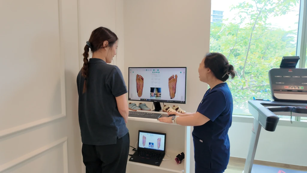 A podiatrist and a patient stand at a clinic workstation reviewing digital scans of both soles on a monitor and laptop, the scanning step used to design custom orthotics.