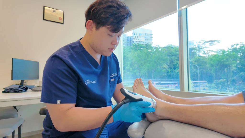 Straits Podiatry 的足科医师身穿深蓝色制服、戴着蓝色手套,在可俯瞰绿意的明亮诊室内,将手持式冲击波治疗探头贴于患者的足跟上。