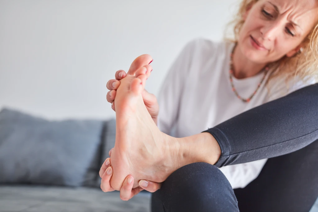 A person seated on a sofa grimaces while cupping the toes and forefoot of one bare foot, the big toe joint region affected in hallux rigidus