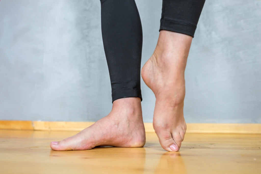 Side view of a person's bare feet on a wooden floor with one heel raised onto the toes, showing the arch profile assessed when checking for high arches.