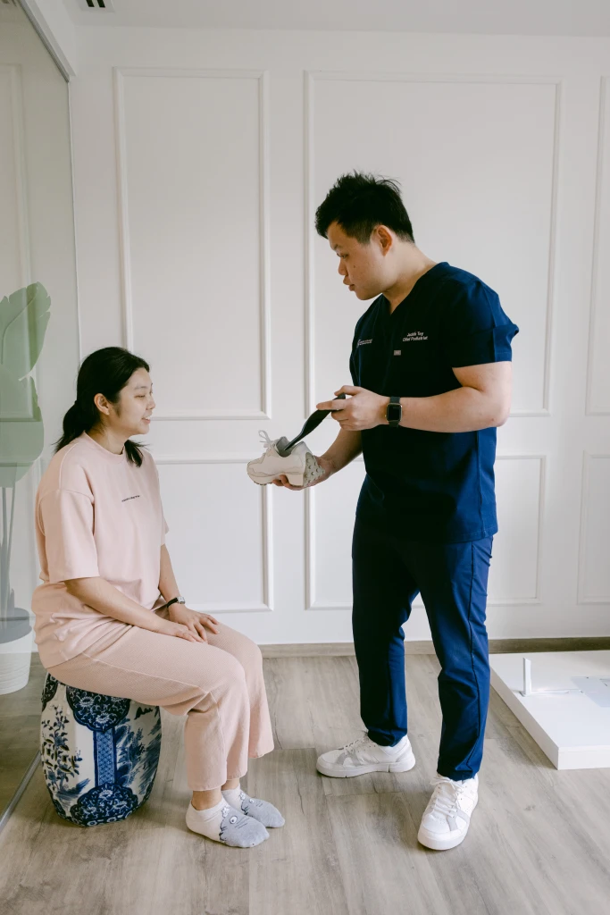 A podiatrist in navy scrubs bending a running shoe to show its flexibility to a seated patient, part of the footwear assessment used when managing high arches.