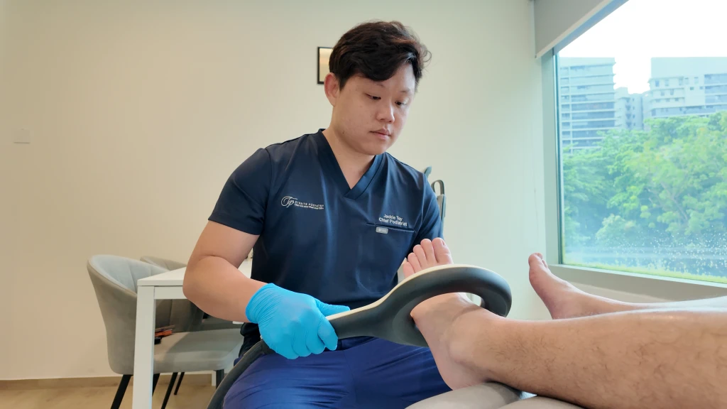 Straits Podiatry 海峡足科一位身穿深蓝色制服、戴蓝色手套的足科医师,将环形磁场传导治疗(EMTT)治疗头置于患者足部与脚踝上方。