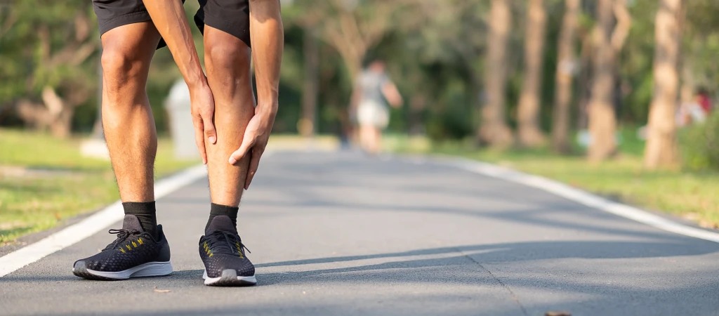 A runner stopped on a park path bending to grip the inner lower shin, the stretch of tibia where shin splints pain is typically felt.