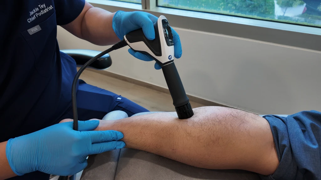 Straits Podiatry 海峡足科的足科医师身穿深蓝色制服并戴着蓝色手套,将手持式径向压力波治疗设备置于患者小腿前侧进行操作。