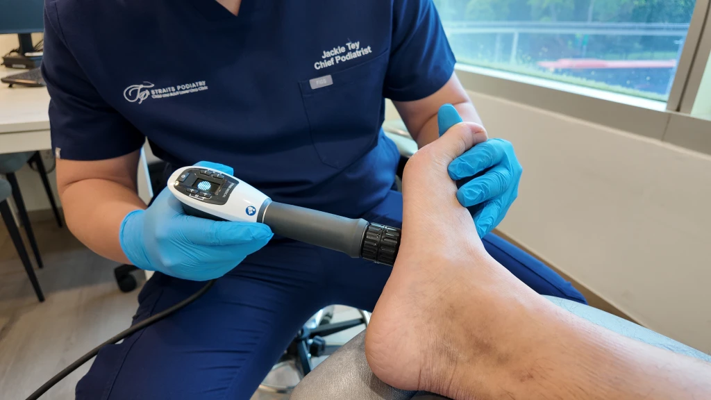 Straits Podiatry 海峡足科的足科医师身穿深蓝色制服并戴着蓝色手套,一手固定患者的足部,一手将径向压力波治疗手柄贴于足跟后侧。