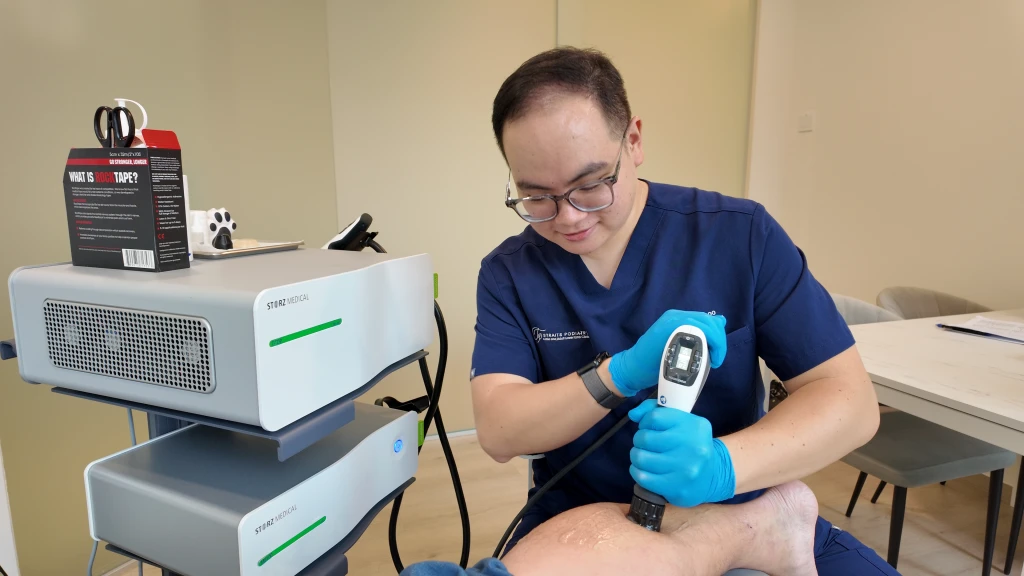 Straits Podiatry 海峡足科的足科医师戴着眼镜、身穿深蓝色制服,将径向压力波治疗手柄用于患者足部,身旁推车上叠放着两台治疗仪。