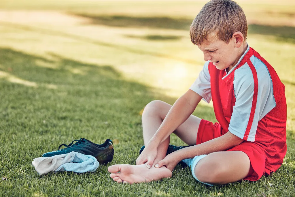 A young footballer in a red kit sitting on grass gripping a bare heel with the boot removed, the growth plate heel pain seen in Sever's disease.