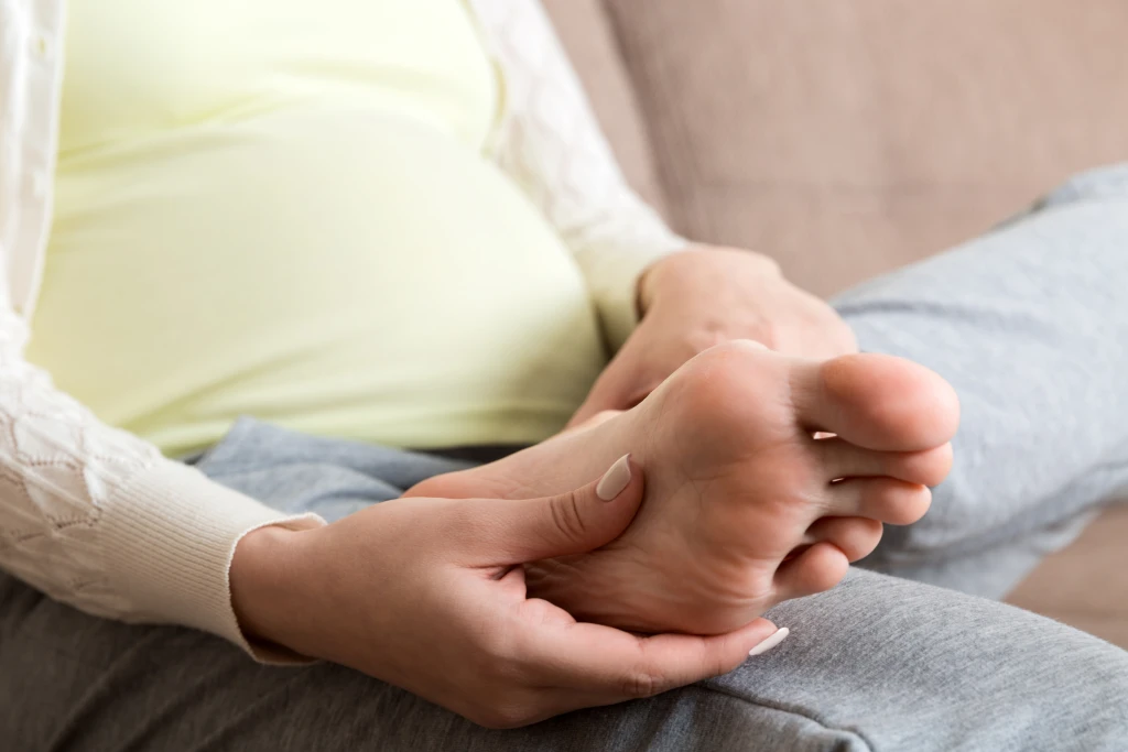 A pregnant person seated and pressing the sole of a bare foot with both hands, pregnancy swelling being one factor that can bring on tarsal tunnel syndrome.