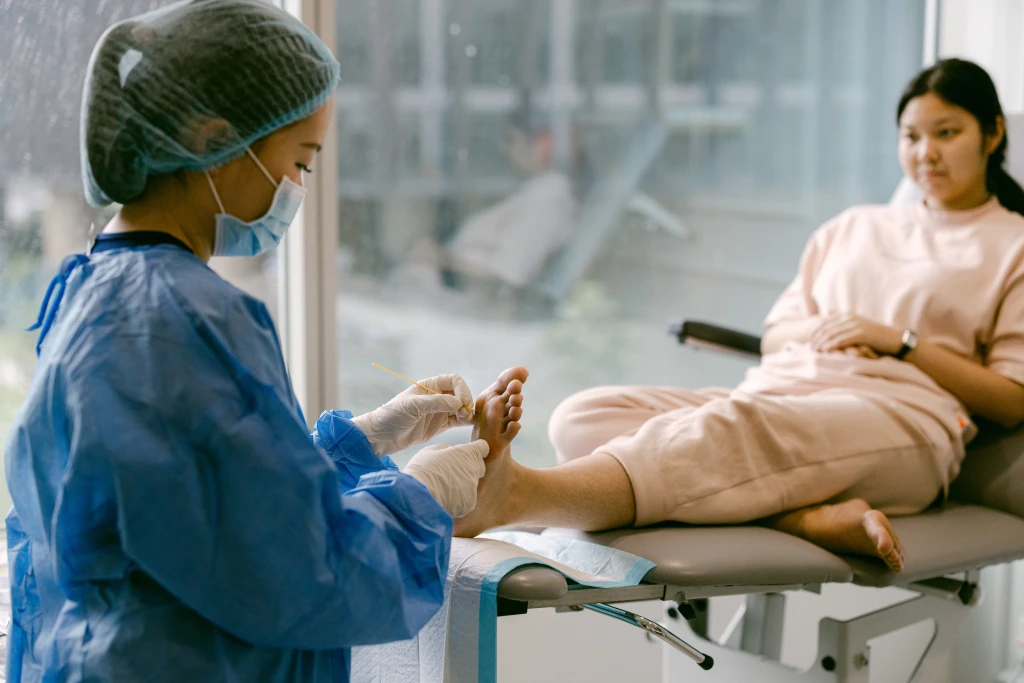 Straits Podiatry 海峡足科的足科医师身穿深蓝色制服并戴着蓝色手套,俯身在窗边用小器械处理患者的足底。