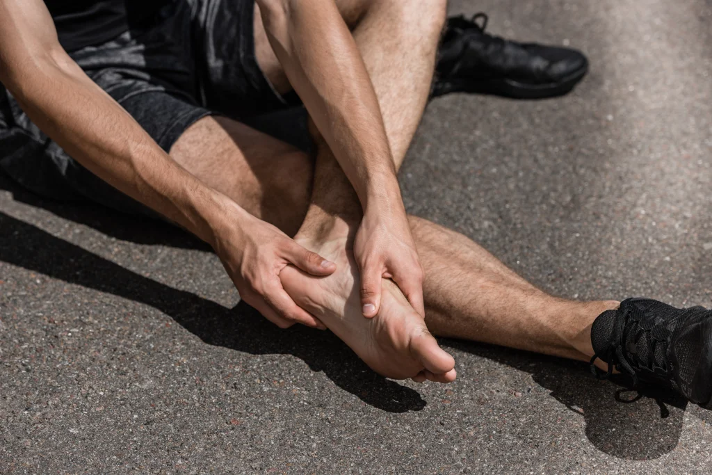 A person in running shoes sits on sunlit asphalt gripping their bare foot and ankle, one of the common foot and ankle injuries that follow sport.
