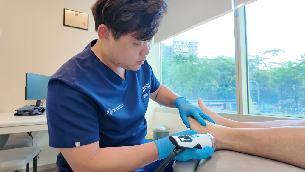 A podiatrist in gloves steadies a patient's foot while applying a shockwave handpiece to the outer ankle, part of peroneal tendonitis recovery before a return to running.