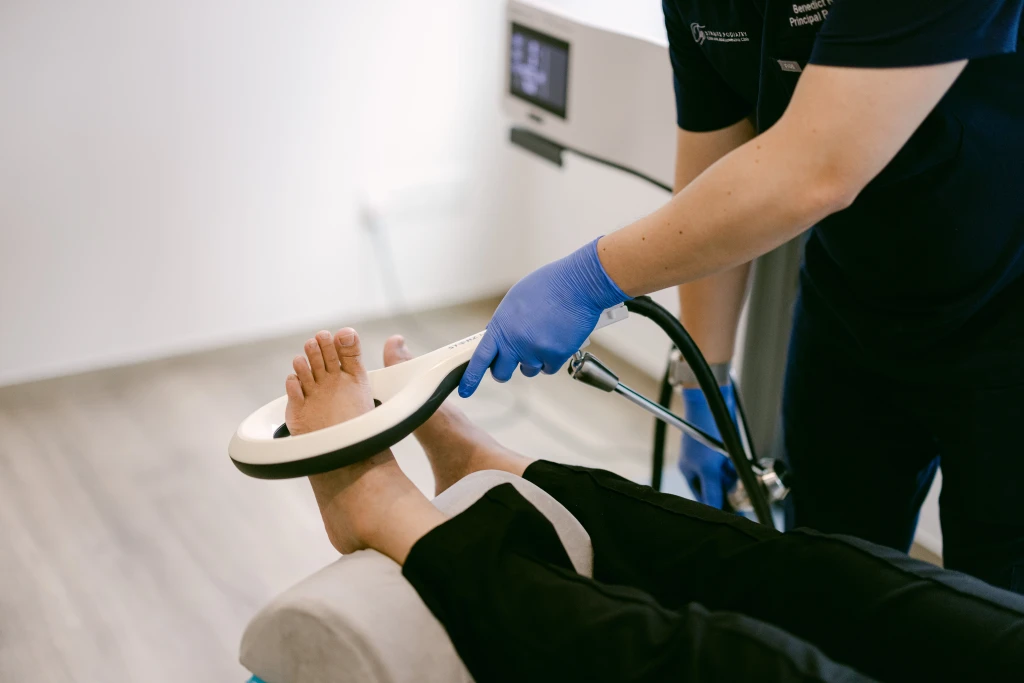 A gloved Straits Podiatry podiatrist positioning a ring-shaped magnetotransduction therapy (EMTT) applicator around a patient's foot and ankle in the treatment room.