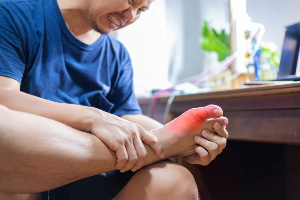 A seated person grimacing and gripping their bare foot, with a red glow marking pain at the big toe joint where turf toe occurs.