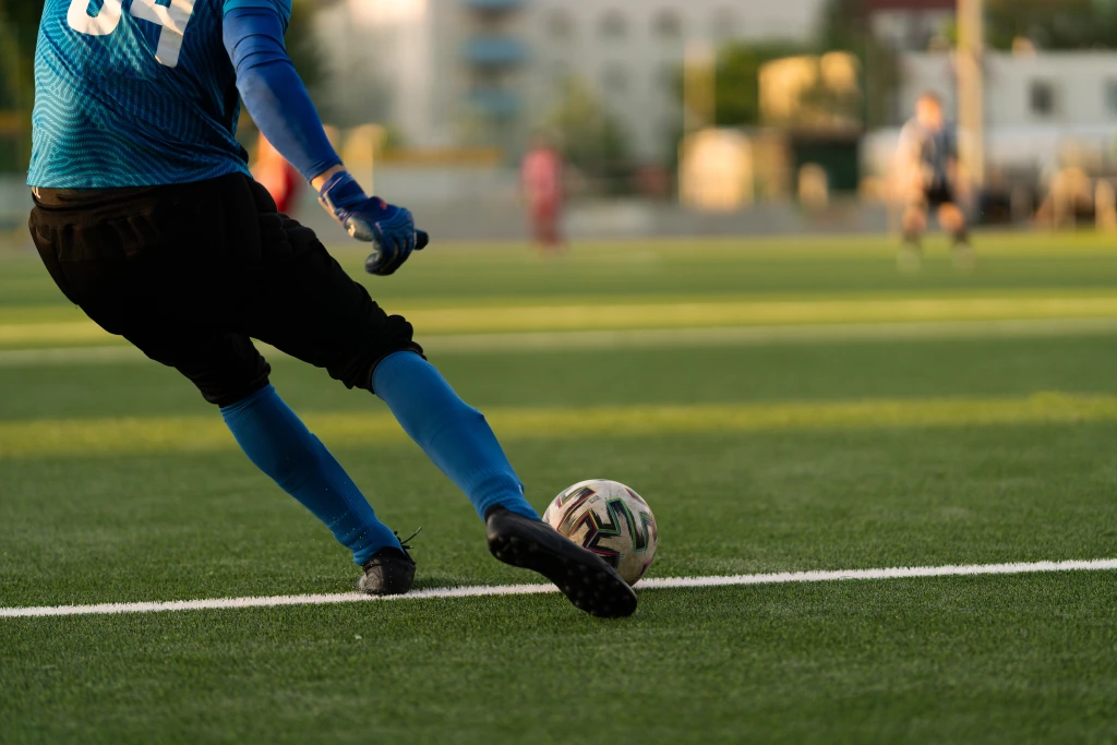 A footballer striking a ball on a green artificial turf pitch, the playing surface associated with turf toe injuries of the big toe.