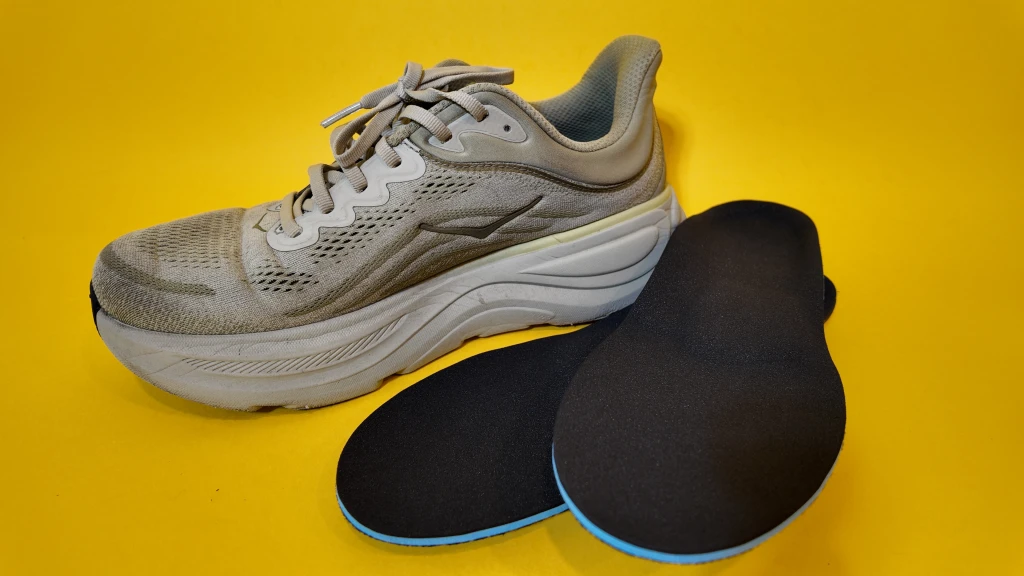 A cushioned mesh running shoe beside a pair of black custom sports orthotics with blue base layers, photographed on a bright yellow background.