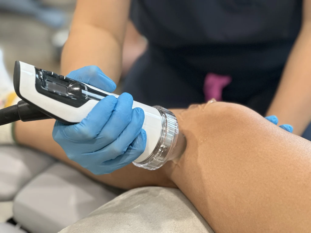 A gloved Straits Podiatry podiatrist applying a handheld focused shockwave therapy (ESWT) device to a patient's knee, with coupling gel over the treatment area.