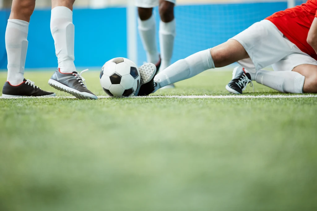 Footballers' legs on a green pitch as one player slides in with an outstretched leg to challenge for the ball, a common mechanism behind football injuries.