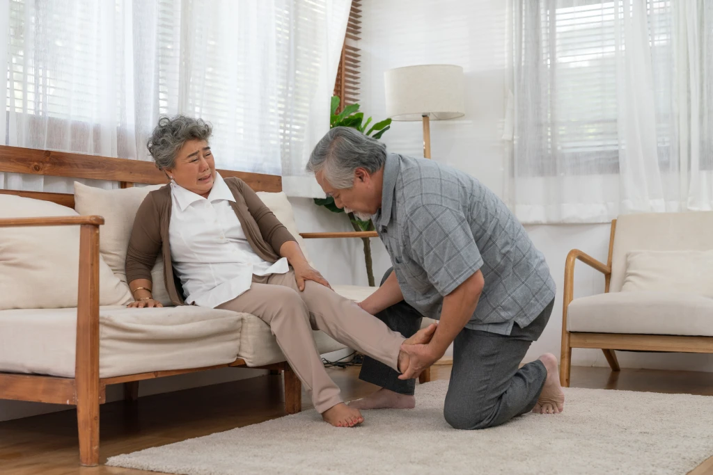 An older adult winces on a sofa while another person kneels and cradles their heel, the heel pain that raises the question of plantar fasciitis versus a heel spur.