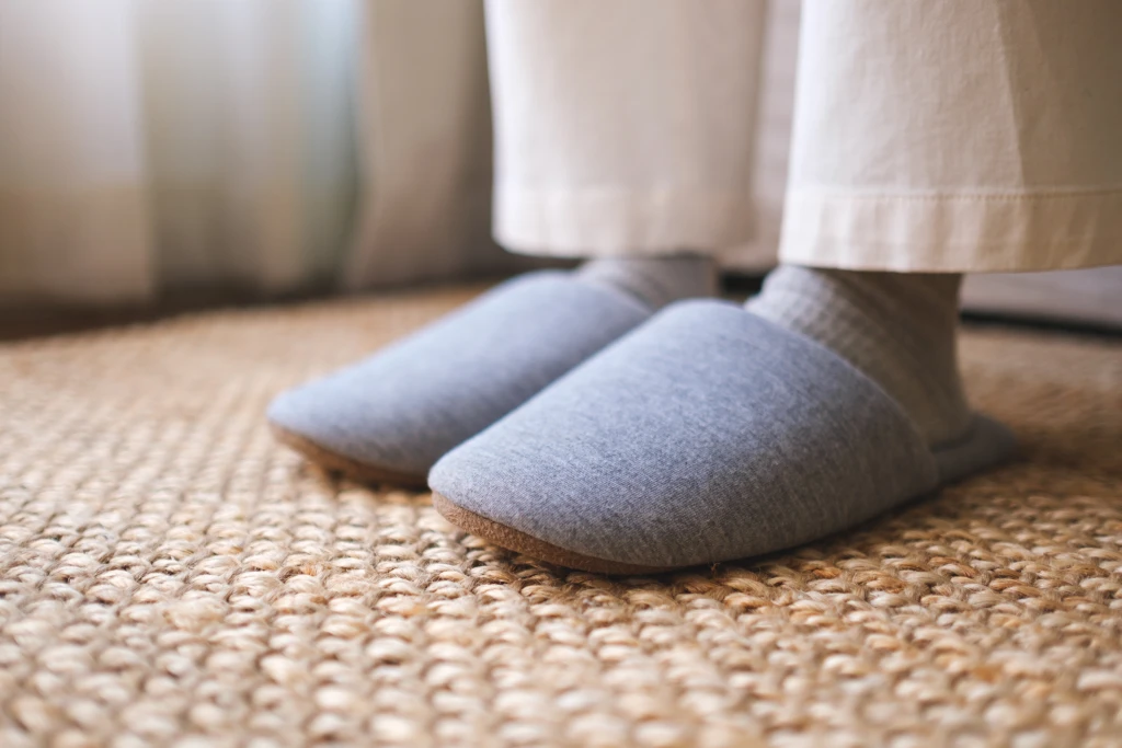 A person in socks on a woven rug wearing soft grey slip-on house slippers with thin flat soles, indoor footwear that gives the arch little support in plantar fasciitis.