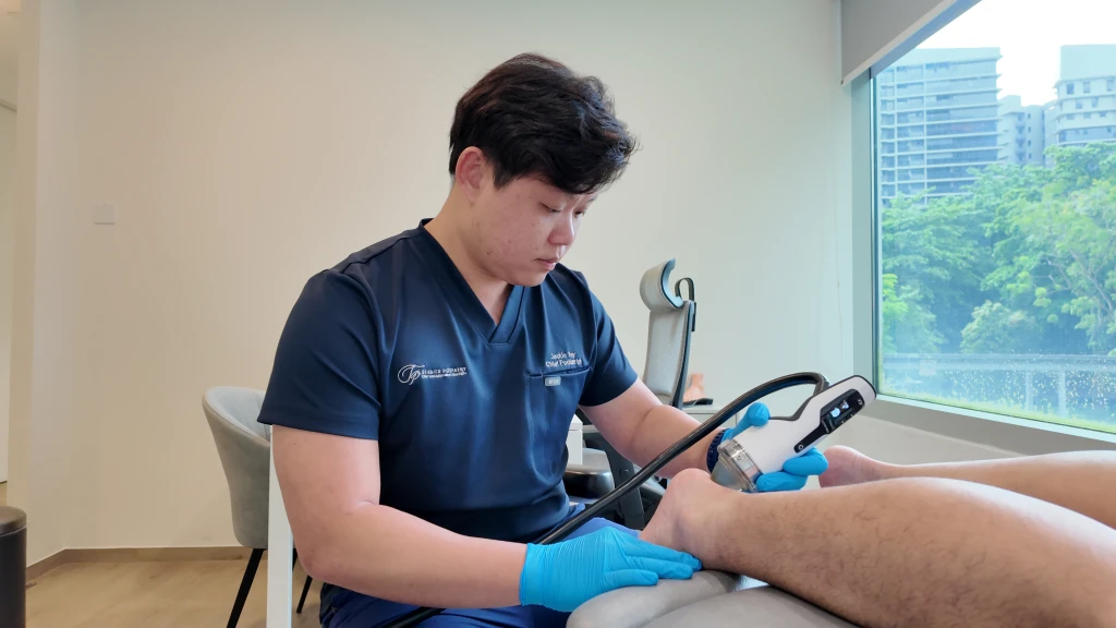 A podiatrist in navy scrubs and blue gloves applying a handheld shockwave device to the back of a patient's lower leg, an option used to manage heel bursitis.