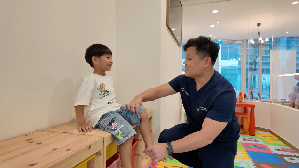 A podiatrist in navy scrubs crouches to talk with a smiling young child seated on a bench in a clinic play area, the paediatric setting where metatarsus adductus is assessed.