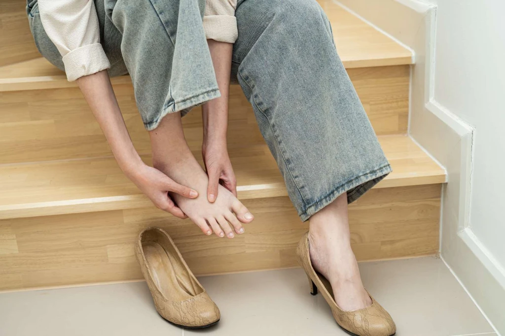 Woman gripping her midfoot after a misstep on wooden stairs, illustrating a common step-off mechanism that can trigger a Lisfranc injury.