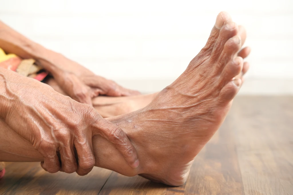 An older person sitting on the floor gripping the ankle of a bare foot, indicating foot and ankle pain