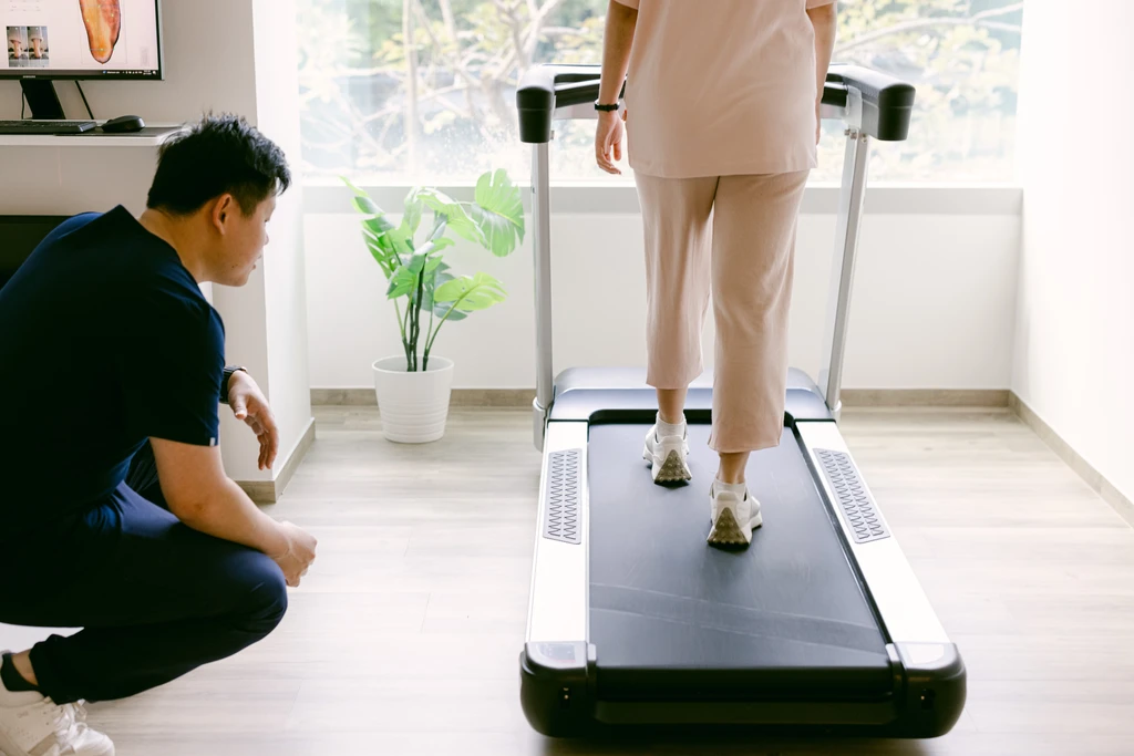 A Straits Podiatry podiatrist observing a patient walk on a treadmill during a gait assessment, with foot-scan imagery on the screen behind.