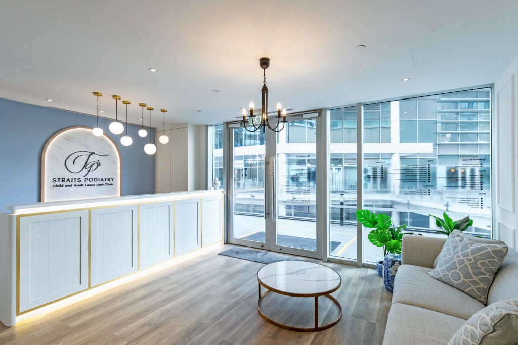 The reception and waiting lounge at a Straits Podiatry clinic, with the Straits Podiatry signage behind the front desk and glass entrance doors.