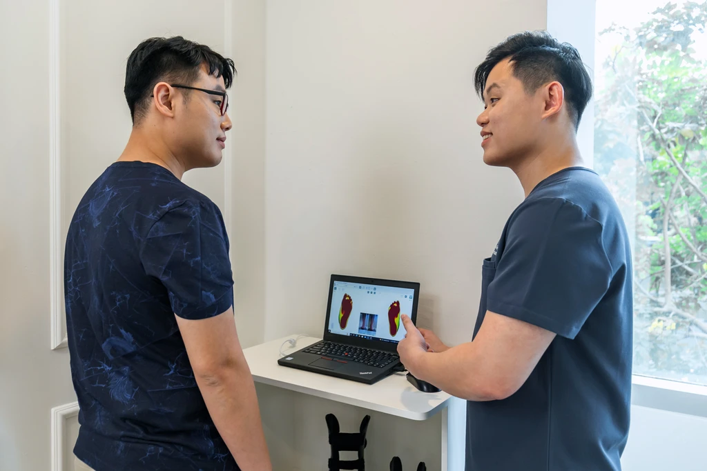 A Straits Podiatry podiatrist explaining a patient's digital foot assessment results on a laptop during a consultation.