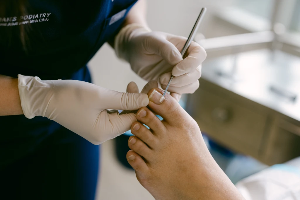 A Straits Podiatry podiatrist in gloves lifting a patient's big toenail with a fine metal instrument during a nail bracing assessment.