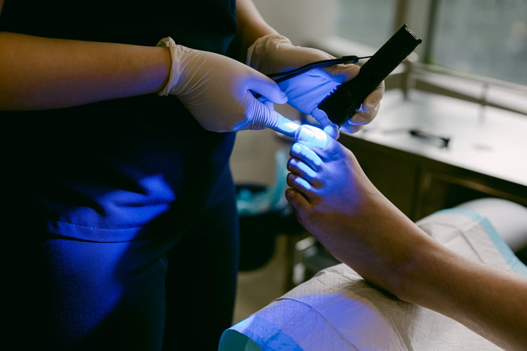 A Straits Podiatry podiatrist applying a handheld curing lamp emitting blue light over a patient's big toenail during a nail bracing procedure.