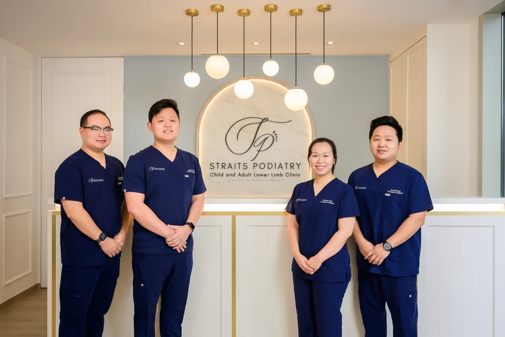 The four podiatrists of Straits Podiatry in navy scrubs at the clinic reception, beneath the Straits Podiatry Child and Adult Lower Limb Clinic signage.