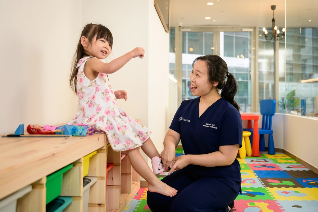 Ms Michelle Goh, Principal Podiatrist, fitting a small foot device for a smiling young girl in the clinic's children's corner.