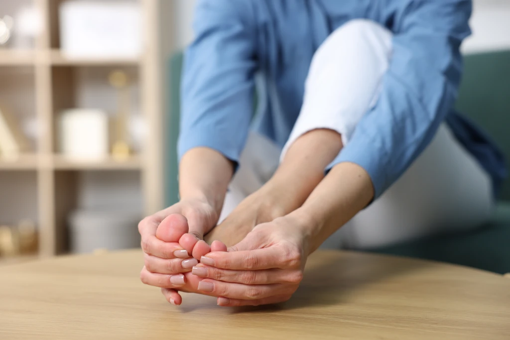 A person seated at home cupping a bare foot with both hands, with reddening across the ball of the foot and toes, a common way toe pain is felt.