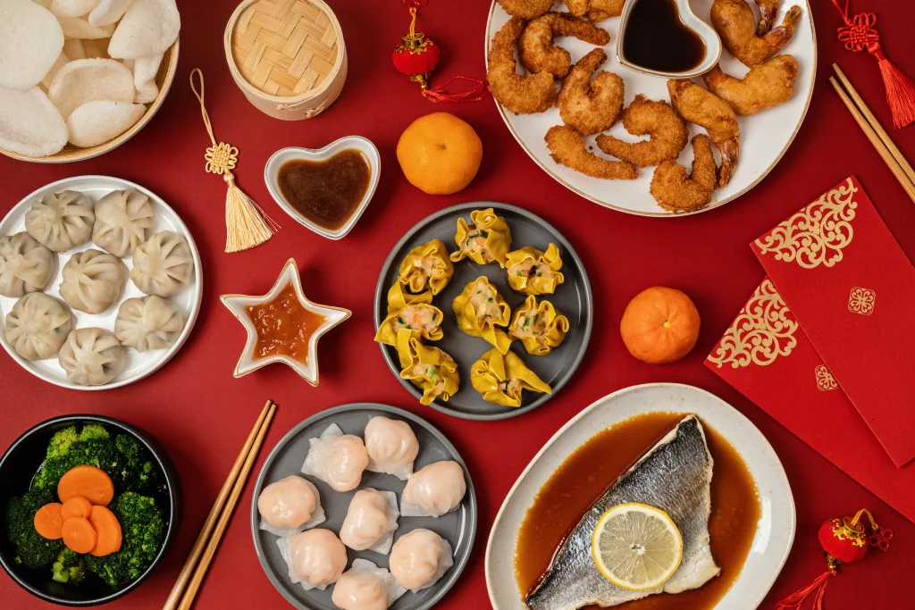 An overhead Chinese New Year spread of fried prawns, dumplings, steamed fish and mandarin oranges on a red background, a table rich in common gout trigger foods.
