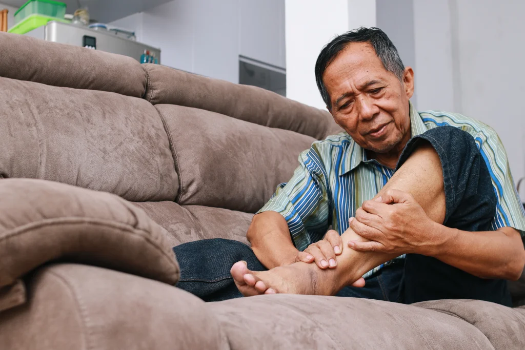 An older person on a home sofa winces while holding a bare foot and ankle, the setting where people learn how to check feet for diabetes.